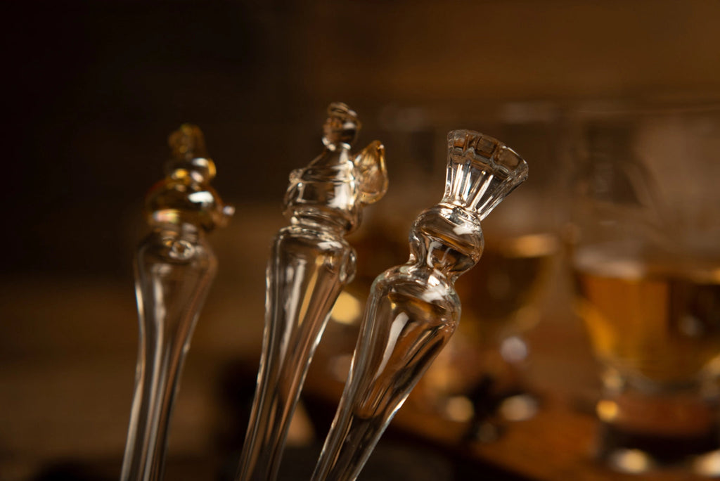 Three glass water droppers shaped like thistle an angel and a potstill with a blurred background of whisky glasses and drinks.